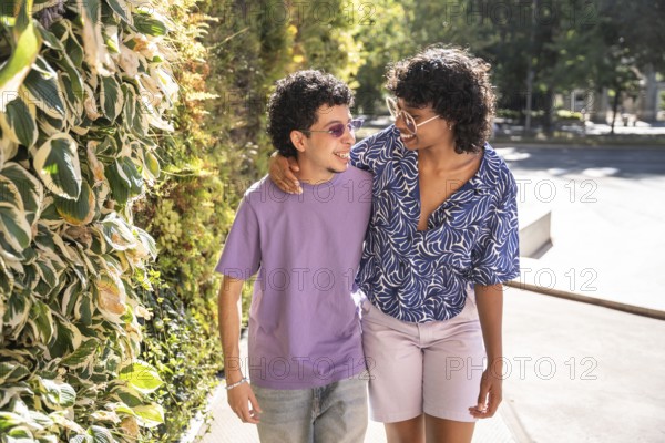 A happy, multiethnic LGBTQ+ couple walking side by side, smiling under the sunlight. Their warm connection is highlighted amidst a natural, leafy backdrop