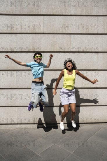 A joyful, multiethnic LGBTQ+ couple, wearing vibrant summer attire, jumps against a concrete wall, symbolizing happiness and diversity on a sunny day