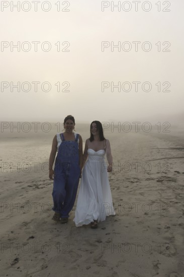 Two women walk along a foggy beach, one in overalls and the other in a flowing white dress The misty atmosphere adds a serene and mystical feel to the scene