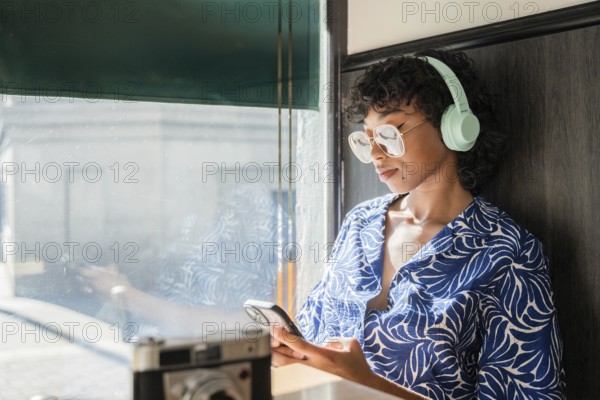 A stylish young woman with curly hair and glasses wearing headphones, leans against a window while engaged with her smartphone. Sunlight filters in, creating a serene ambiance