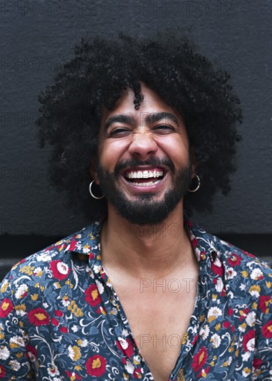 A man with curly hair laughs joyfully, wearing a vibrant floral shirt against a gray background. His expression radiates happiness and positivity, capturing a moment of pure joy