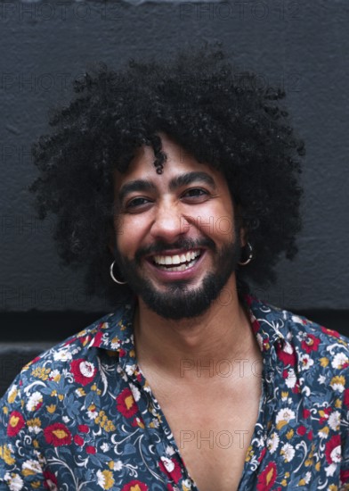 A joyful man with curly hair and hoop earrings smiles against a gray background. Wearing a vibrant floral shirt, they radiate warmth and confidence in a casual setting