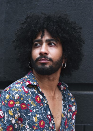 Portrait of a stylish man with curly hair wearing hoop earrings and a vibrant floral shirt. The image captures modern fashion and urban style against a gray background