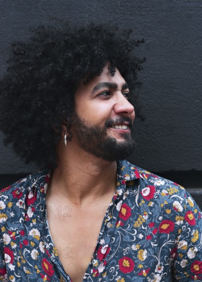 A man with curly hair and a beard smiles while wearing a vibrant floral shirt against a gray background. His joyful expression reflects a casual style and confident demeanor