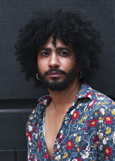 A close up portrait of a man with curly hair, wearing a floral shirt and hoop earrings against a gray background. The expression is serious and contemplative