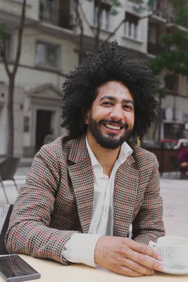 A cheerful Mixed-race man with curly hair teleworking from a street-side cafe, using a laptop and enjoying a cup of coffee