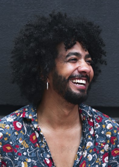 A smiling man with curly hair and a floral shirt exudes happiness. Their joyful expression and casual attire create a lively and warm atmosphere a gray background