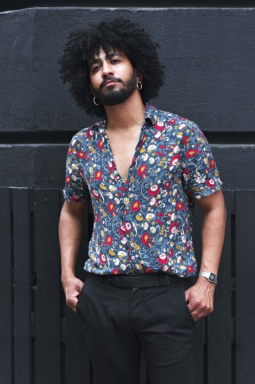 A stylish individual with curly hair and beard leans against a gray wall, wearing a vibrant floral shirt and dark trousers, exuding confidence and modern fashion