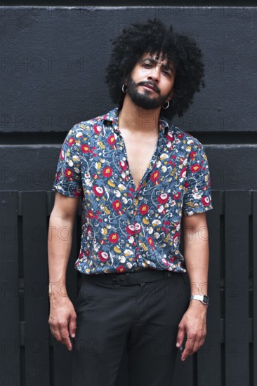 A confident young man with curly hair poses in a vibrant floral shirt against a gray urban wall, showcasing modern fashion and a relaxed, stylish demeanor