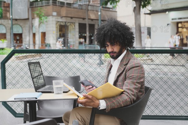 A Mixed-race man with curly hair is engaged in remote work using a laptop and smartphone at an outdoor cafe, embodying modern telework flexibility