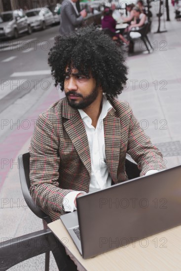 A Mixed-race man with curly hair, dressed in a stylish plaid jacket and white shirt, is focused on his work from a laptop outdoors in a vibrant urban street setting