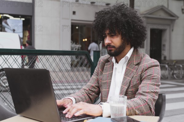 A mixed-race male with curly hair is teleworking at an outdoor cafe in a bustling city environment, focused on his laptop