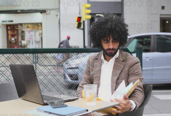 A young Mixed-race man with curly hair working remotely at a street-side cafe, carefully reviewing documents while surrounded by urban life