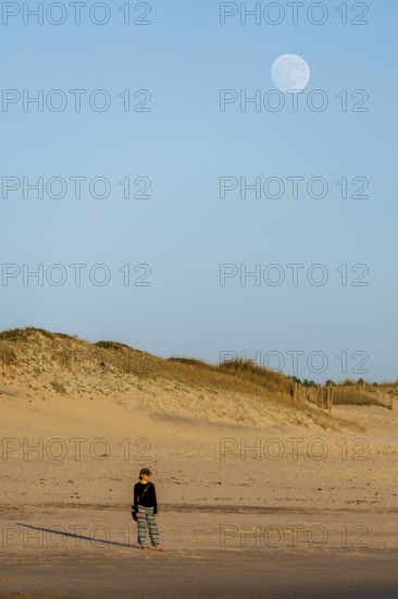 A peaceful scene of a woman standing on the sandy beach at twilight, with a large moon rising above the dunes in Vila do Conde, Portugal