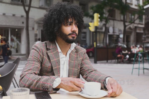 A Mixed-race man with curly hair works remotely, utilizing digital devices at a street cafe while sipping coffee
