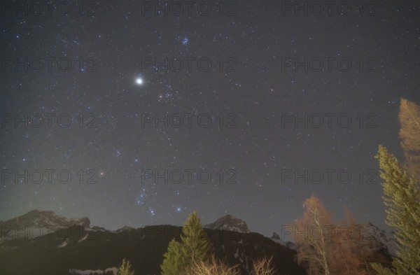 The constellations of Orion and Taurus shine brightly in the night sky, accompanied by Jupiter, Sirius, the brightest star, and the Pleiades. Snowy alpine peaks and trees enhance the celestial view