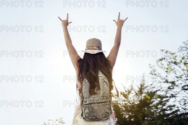 A woman wearing a hat and backpack raises her arms in a joyful sign, celebrating freedom and nature. The bright sky and greenery create a vibrant, uplifting scene