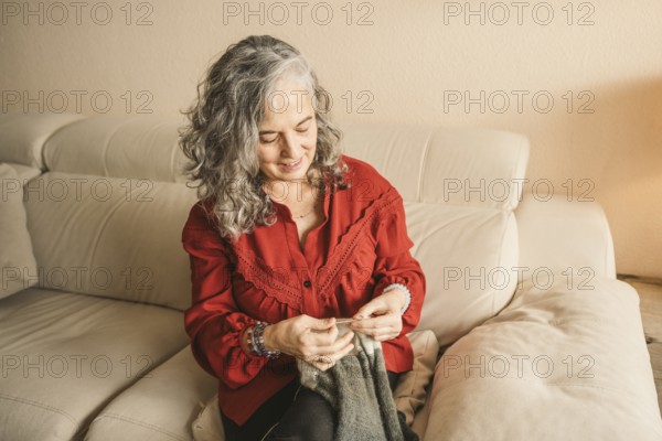 A woman with gray hair sits comfortably on a beige sofa, focused on knitting. She wears a red blouse, conveying warmth and creativity in a cozy environment