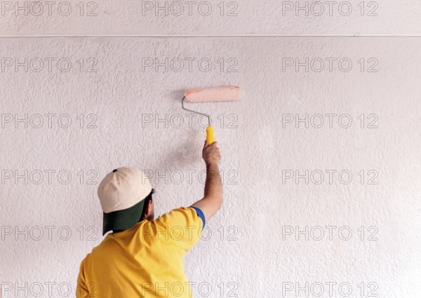 Back view of unrecognizable man in yellow shirt painting a white wall with a roller. This image captures a moment of home remodeling work, focusing on wall decoration