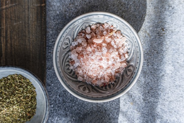 Close-up of Himalayan pink salt in a decorative ceramic bowl alongside fresh green herbs on a rustic wooden and stone background, perfect for healthy cooking inspiration