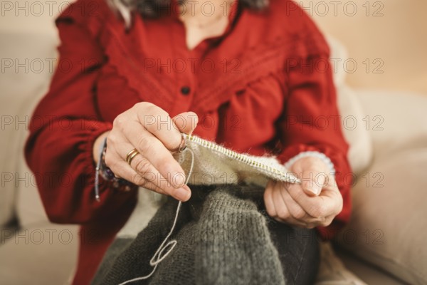 A woman in a red blouse sits comfortably on a couch, skillfully knitting a wool sweater. The close-up focuses on her hands engaged in the craft, emphasizing the texture and details