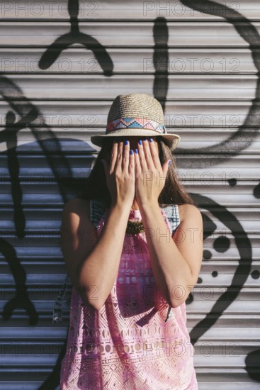 A woman in a straw hat and pink dress stands against a graffiti wall, covering her face with her hands, showing off her blue nail polish, creating a playful and stylish scene