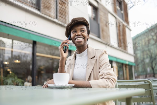Black woman sits at an outdoor cafe, smiling and talking on her smartphone She is wearing a stylish outfit, enjoying her coffee Perfect depiction of urban lifestyle