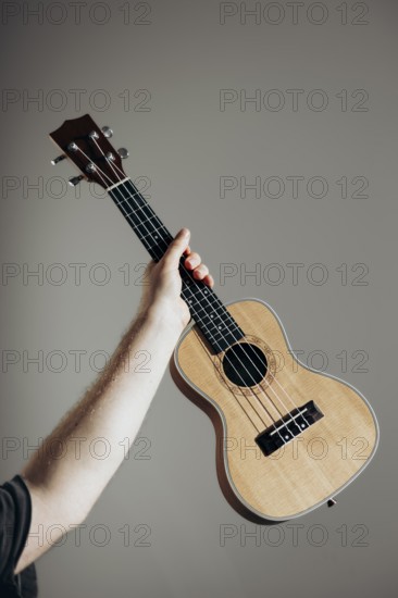 A person holds a wooden ukulele aloft against a neutral background The lighting highlights the instrument's natural finish, creating a serene and artistic composition