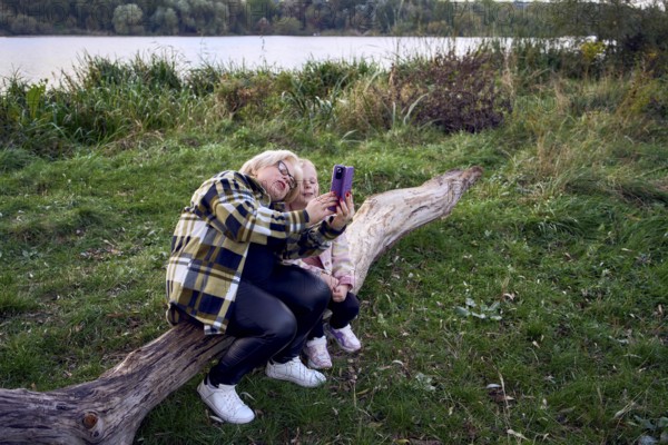 A mother and daughter sit on a wooden log by a scenic riverside, capturing a joyful selfie together. The lush greenery and tranquil water create a serene backdrop