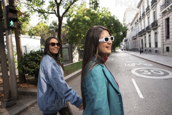 A joyful Latin lesbian couple strolls hand in hand down a tree-lined city street. They wear stylish jackets and sunglasses, embracing their love openly under the sun