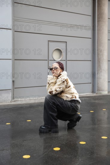 A fashionable woman wearing a fur jacket and dark jeans poses stylishly outdoors, surrounded by a minimalist urban backdrop. Their confident expression and stylish eyewear stand out