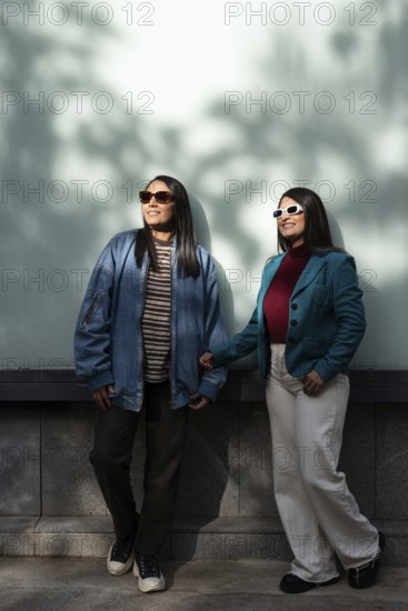 A stylish Latin lesbian couple enjoys a sunny day, dressed in casual fashion with jackets and sunglasses. They stand close, holding hands, casting shadows on the wall behind them