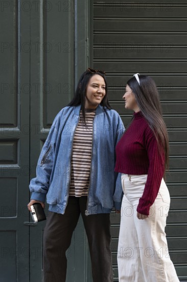 A Latin lesbian couple shares a joyful moment outdoors. One wears a denim jacket and striped shirt, the other a red turtleneck. They gaze at each other with smiles, expressing connection