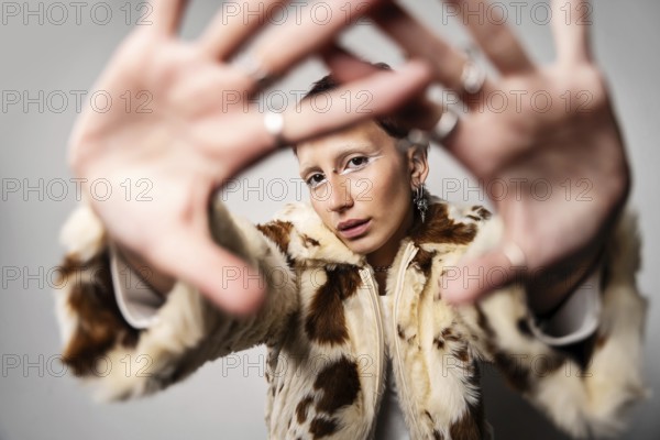 A woman wearing a stylish fur coat poses with hands stretched toward the camera. They sport bold makeup and multiple piercings, creating an edgy fashion statement