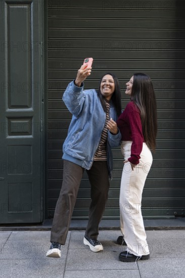 A joyful latin lesbian couple poses for a selfie in an urban setting, showcasing their unity and happiness. The relaxed atmosphere conveys warmth and a strong connection between them