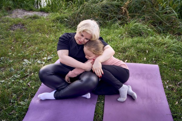 A mother and his daughter share a warm, comforting hug while sitting on yoga mats in a riverside. The moment reflects love, connection, and nature's serenity