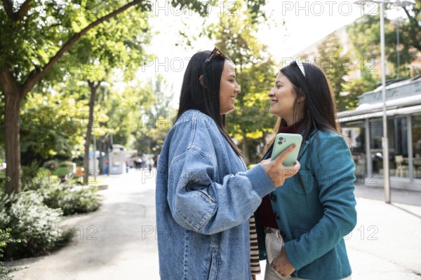A cheerful Latin lesbian couple stands close outdoors, sharing a joyful moment. They smile warmly at each other, surrounded by trees and sunlight, embodying love and happiness