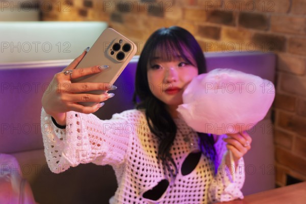An Asian Gen Z girl captures a selfie with her phone while enjoying cotton candy in a vibrant arcade setting, blending fun and modern youth culture