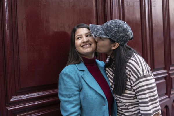 A loving Latin lesbian couple shares a sweet moment by a burgundy wall. One partner playfully kisses the other on the cheek, radiating affection and happiness