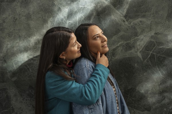 A Latin lesbian couple shares a loving embrace, expressing warmth and affection. The tender moment is captured against a textured background, highlighting their connection