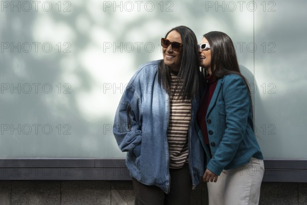 A Latin lesbian couple stands close, basking in the sun. One wears a denim jacket, the other a teal blazer, both sporting sunglasses. The backdrop is a soft, shadowed wall