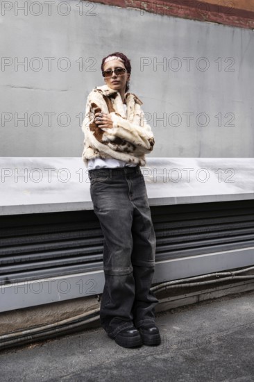 A woman confidently poses in a trendy fur jacket and sunglasses, embodying urban street style. Their casual attire includes wide-leg jeans and platform shoes