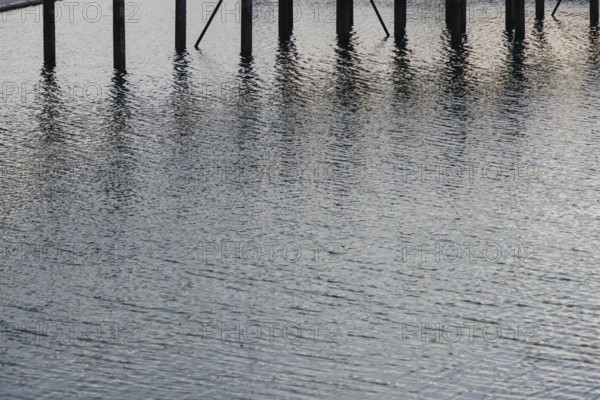 Water reflections with wooden poles standing vertically. The ripples and light reflections create a calm and textured scene