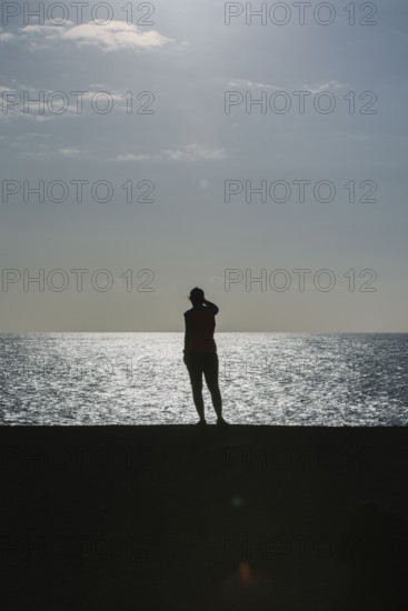 Silhouette of a person standing by the sea during sunset. The calm water and warm light create a peaceful atmosphere
