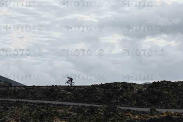 A lone cyclist riding on a volcanic road under a cloudy sky. The contrast between the dark terrain and the clouds creates a dramatic scene