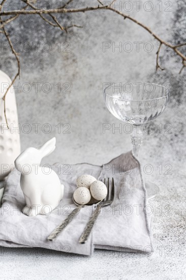 An elegant Easter-themed display featuring a white ceramic rabbit, three speckled eggs, and a clear glass. This setup is complemented by silverware and a textured napkin, presenting a serene holiday mood
