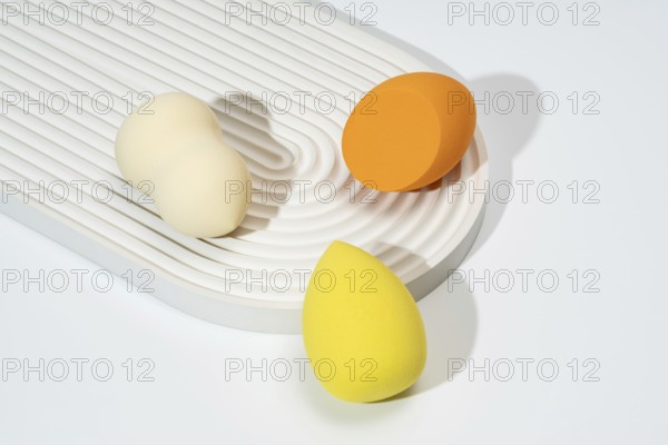 A trio of colorful cosmetic sponges in orange, yellow, and cream are arranged on a textured, white surface The minimalist composition highlights the beauty tools in soft, natural lighting