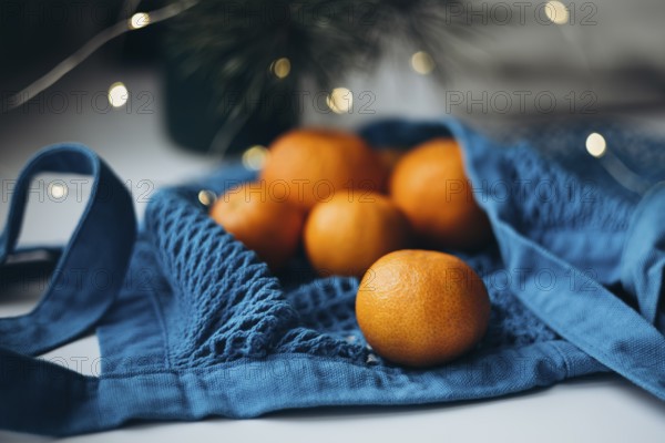 Fresh tangerines placed inside a vibrant blue string bag, set against a softly lit holiday backdrop, create a festive and cozy feel. The blue bag adds a pop of color, enhancing the orange hues of the tangerines