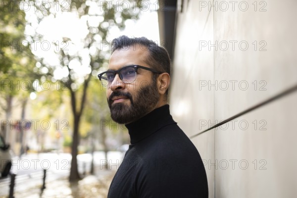Side view of an Indian man dressed in a black turtleneck stands thoughtfully on a city street lined with autumn trees, looking away from the camera