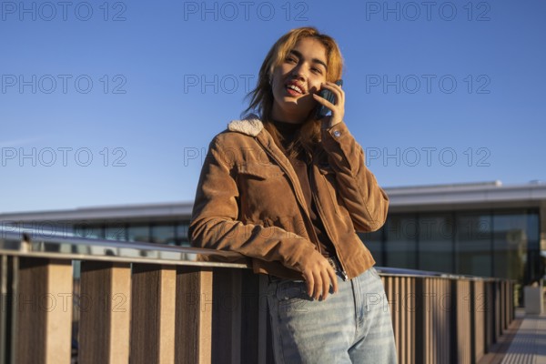 Mixed-race woman smiling while leaning on a railing and talking on a mobile phone outdoors She wears a brown jacket and jeans, with a clear blue sky in the background